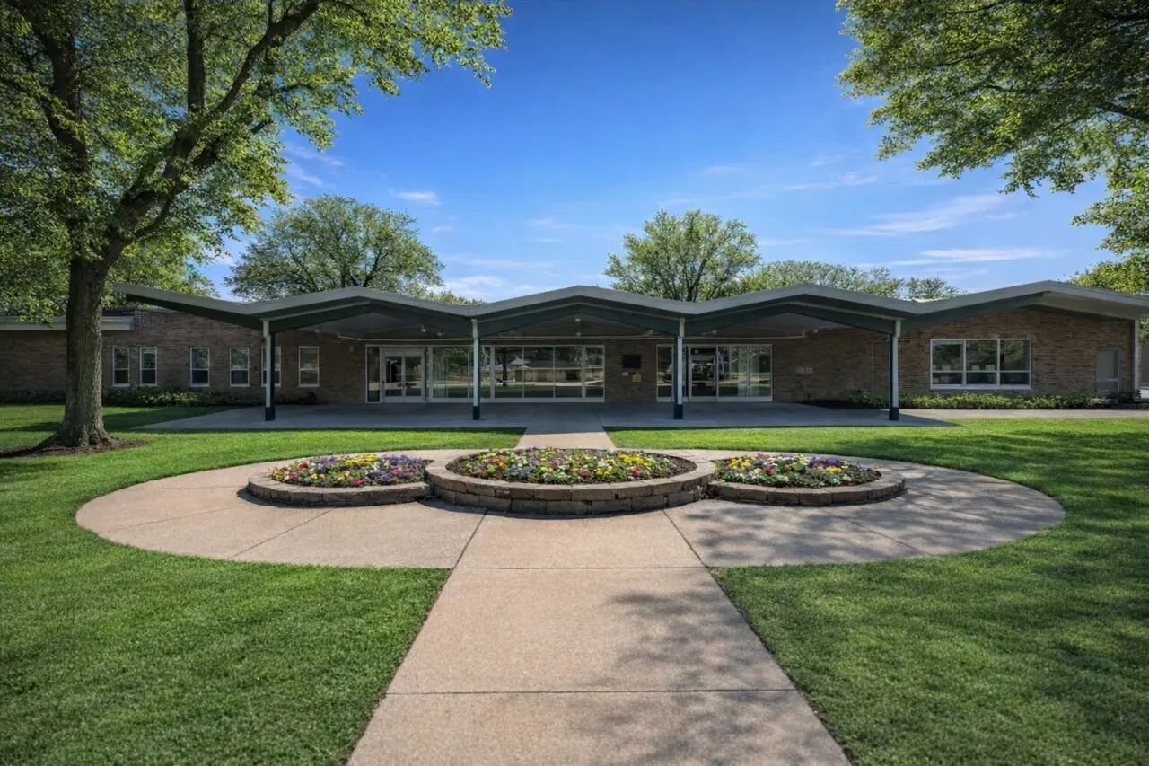 A single-story brick healthcare building with a unique zigzag roofline, surrounded by green grass and trees, with a paved walkway leading to the entrance and three circular flower beds with colorful flowers in front.