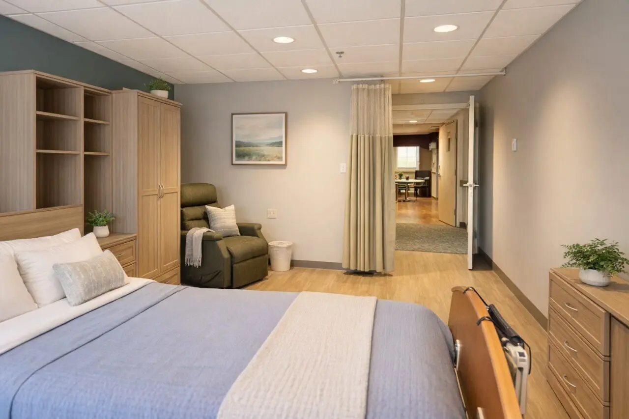 A hospital room with a bed on the left side, topped with white pillows and a beige decorative pillow. There are wooden cabinets and shelves behind the bed, with small potted plants on top. In front of the window, an olive-green recliner chair with a white throw blanket and pillow. A curtain hangs in the doorway leading to a hallway, which opens into another room with a desk and chair. The room has a light-colored wooden floor, gray walls, and a ceiling with recessed lighting.