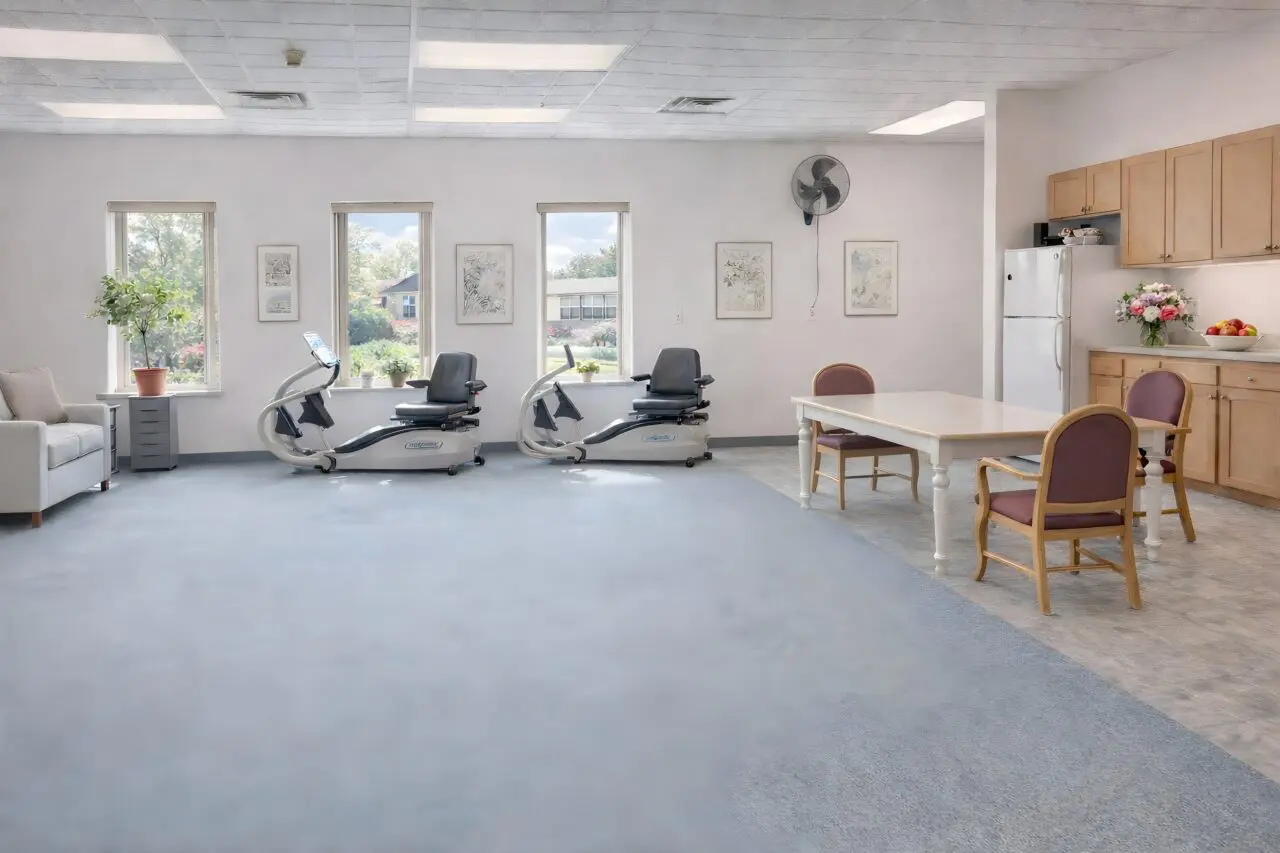 A bright, spacious common area with three large windows, two exercise chairs, a small sofa with a potted plant, a dining table with three chairs, a refrigerator, and kitchen cabinets.