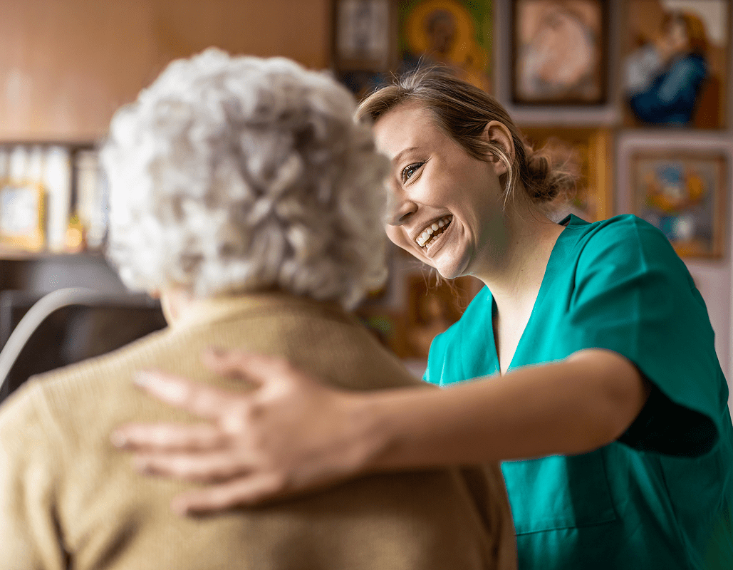 A healthcare worker with a bright smile is embracing an elderly patient, providing comfort and support in a warmly decorated room.