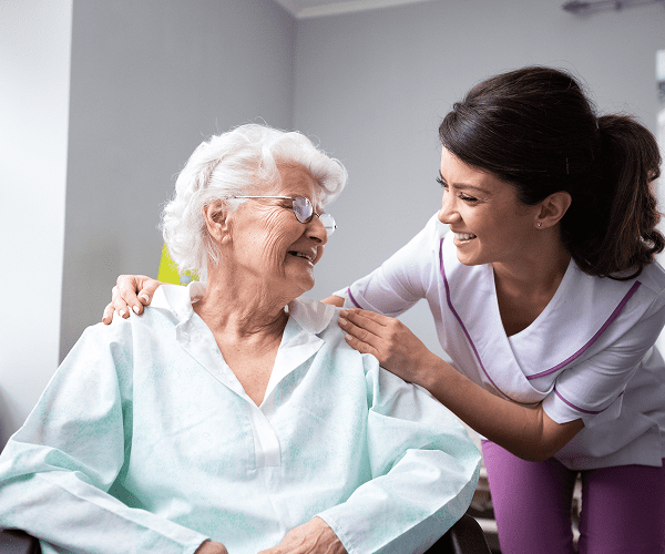 A healthcare worker smiling warmly and leaning toward an elderly woman with white hair and glasses, who is sitting in a hospital gown; the healthcare worker has her hand gently on the woman's shoulder.