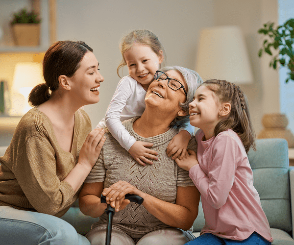 Four generations of women, including an elderly woman with a walker, are happily smiling and embracing each other in a cozy living room.