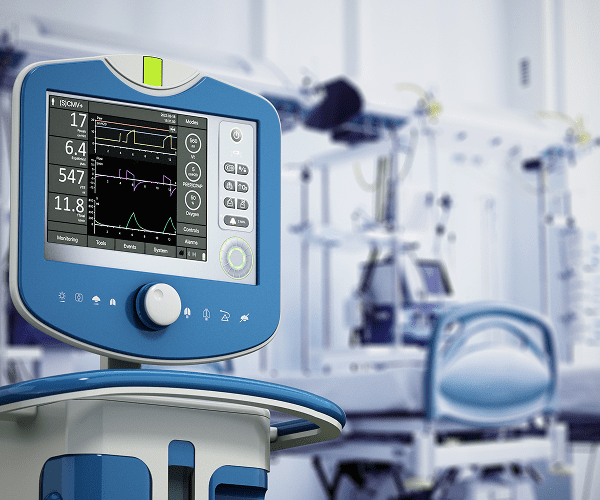 A ventilator machine with a digital display showing vital signs and respiratory data, situated in a hospital room with medical equipment and a hospital bed in the background.