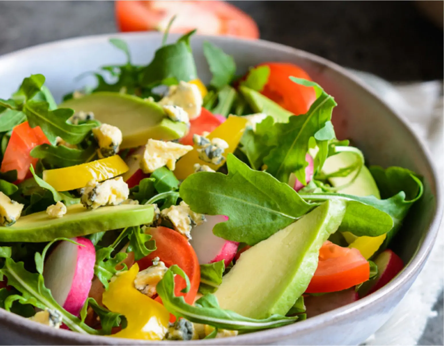 A colorful mixed salad in a white bowl, containing arugula, sliced cucumbers, cherry tomatoes, yellow bell peppers, radishes, crumbled blue cheese, and avocado slices.