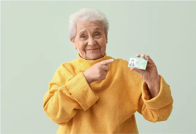 An elderly woman with white hair, wearing a yellow sweater, smiling and pointing to a driver's license she is holding up.