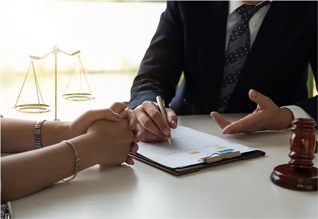 A person in a suit and tie is sitting at a desk, signing a document with a pen, while another person holds their hands. A gold gavel and a set of scales are positioned on the desk, symbolizing legal or judicial themes.