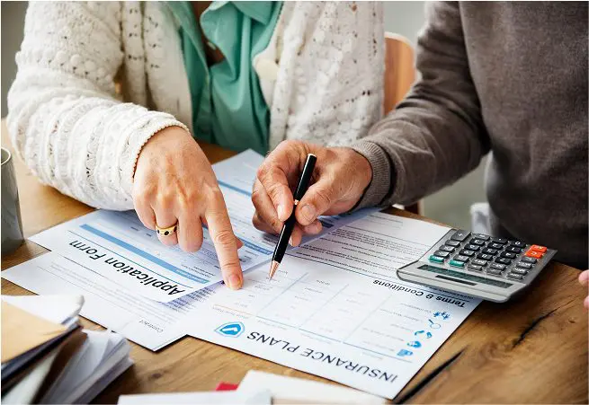Two people reviewing insurance documents at a table; one is pointing to a form with a finger, the other is holding a pen, with a calculator nearby.
