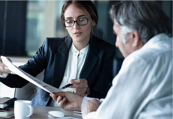 A young woman in glasses and a dark blazer discusses documents with an older man in a white shirt at a table in an office setting.