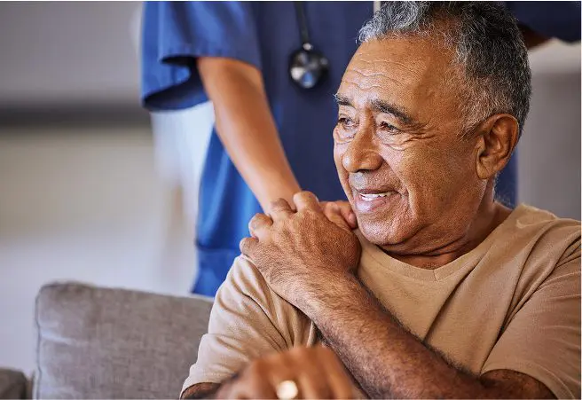 A smiling elderly man sitting on a couch with a healthcare worker standing behind him, gently touching his shoulder. The healthcare worker is wearing blue scrubs and a stethoscope.