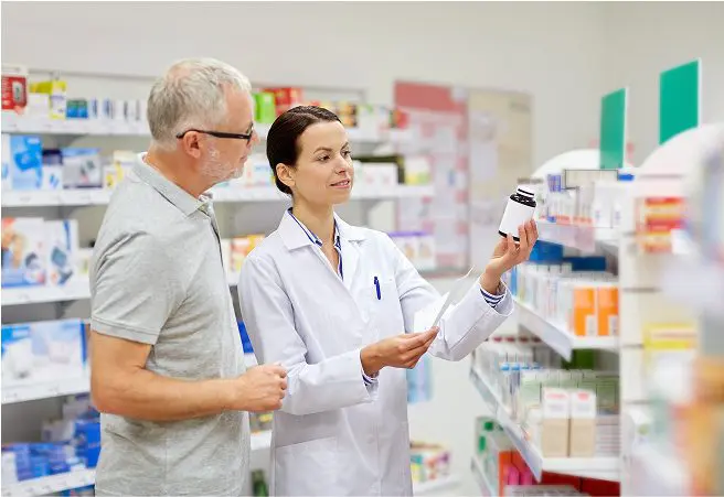 A female pharmacist wearing a white lab coat shows a medication bottle to a male customer with gray hair and glasses in a pharmacy aisle filled with various medicine boxes and bottles.