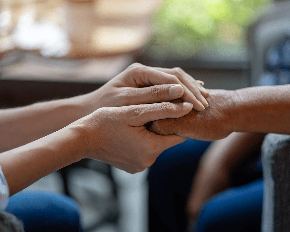 A person gently holding and comforting an elderly person's hand inside a healthcare setting.